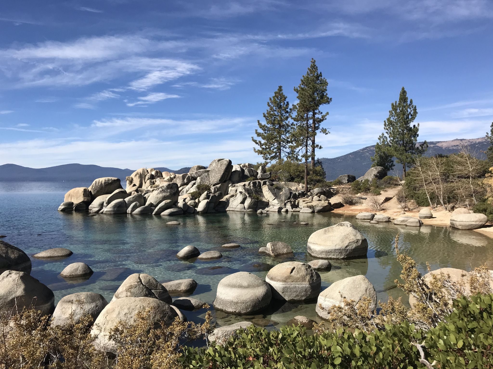 Smooth granite boulders in the clear turquoise shallows of Lake Tahoe under a blue sky with pines. Ulana Rey PharmD