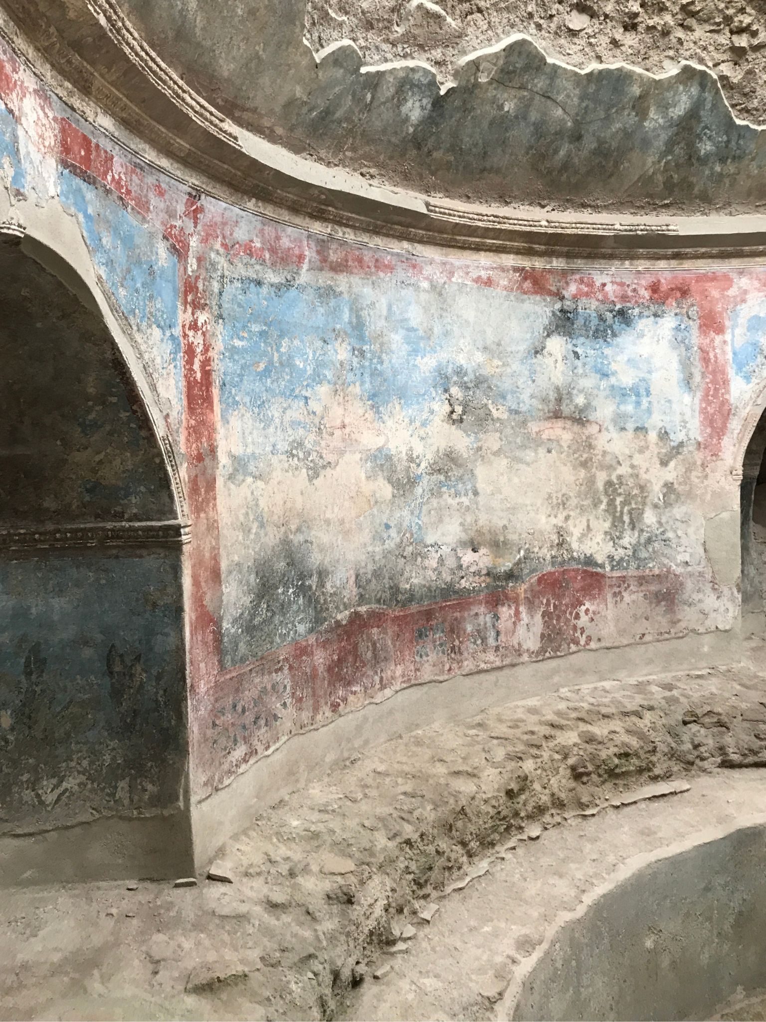 Weathered red and blue frescoes on the curved plaster walls of an ancient Roman bath at Pompeii. Ulana Rey PharmD